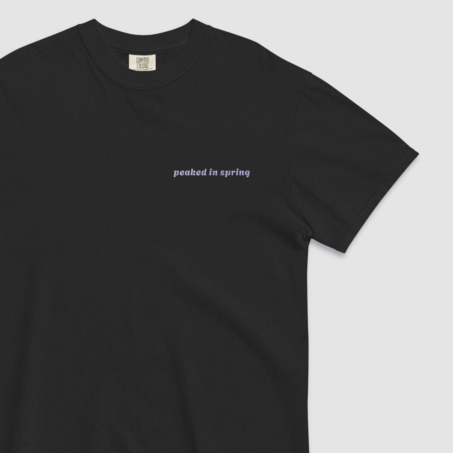 Close up of front of black tshirt with 'peaked in spring' written on the left chest in lilac