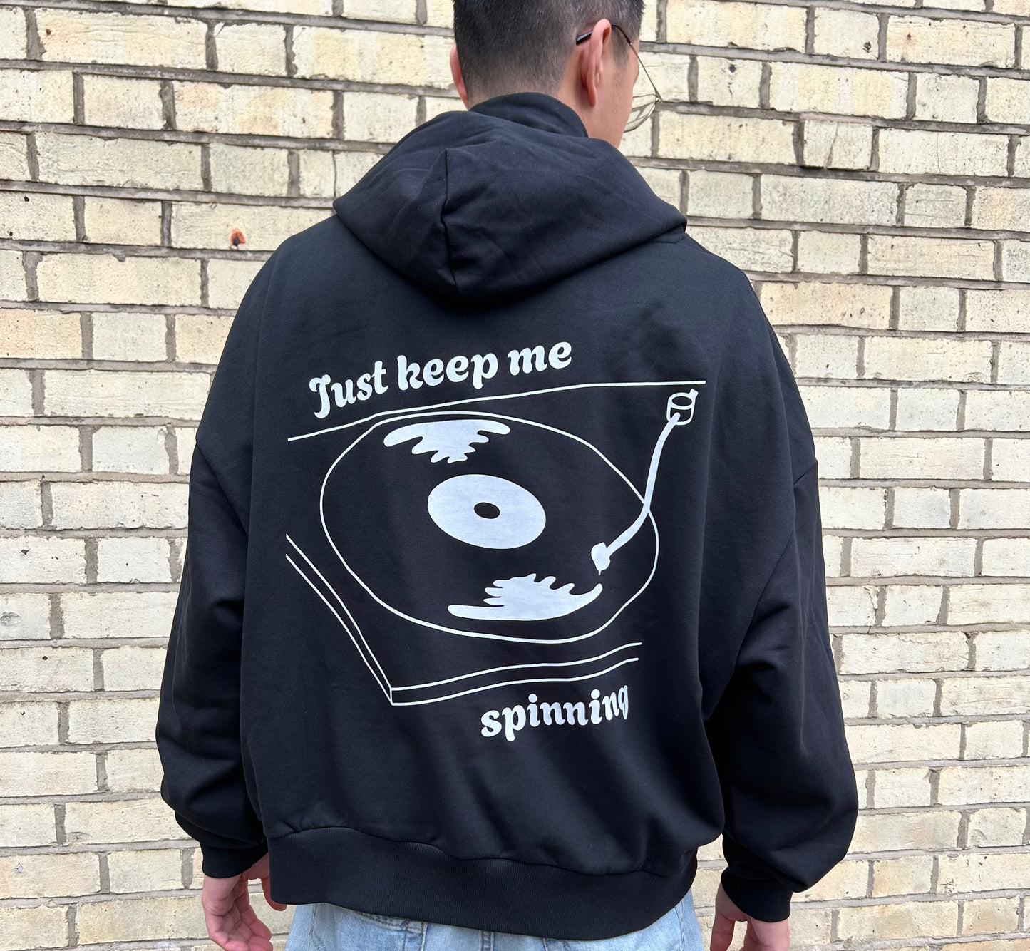 Person wearing a black hoodie with a vinyl graphic and text on the back, standing against a brick wall.