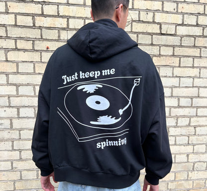 Person wearing a black hoodie with a vinyl graphic and text on the back, standing against a brick wall.
